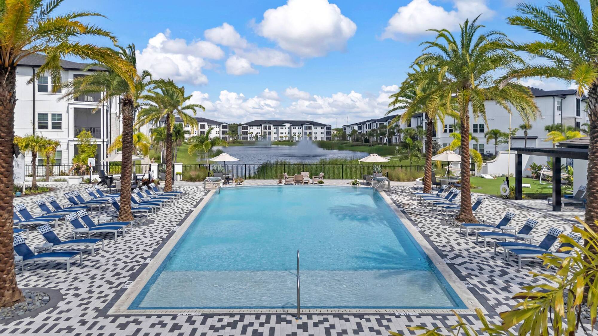 Lakeside resort pool at Avasa Hammock Landing in West Melbourne, FL with palm trees, umbrellas, and apartment buildings beyond.